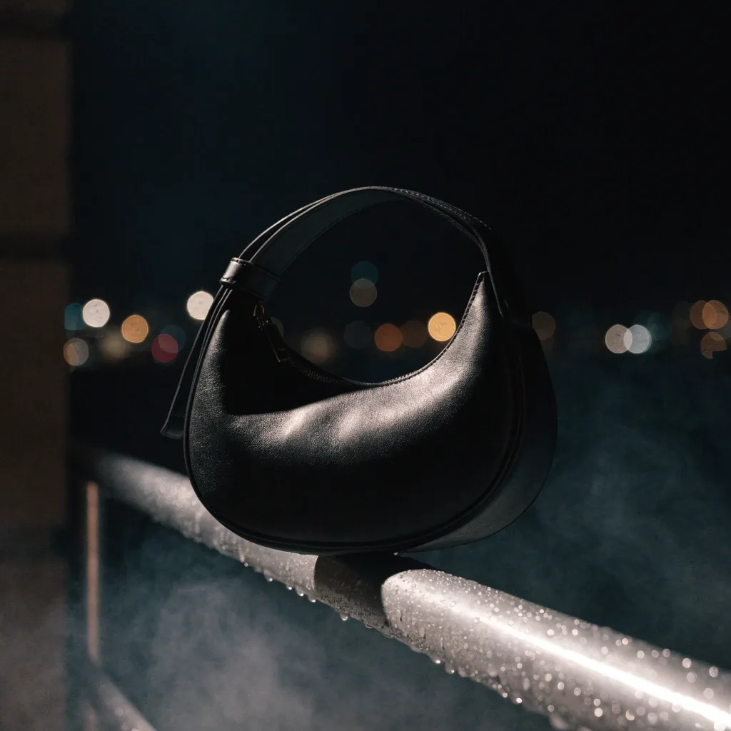 A crescent crossbody bag resting on a fogged metal railing at night with city lights far behind