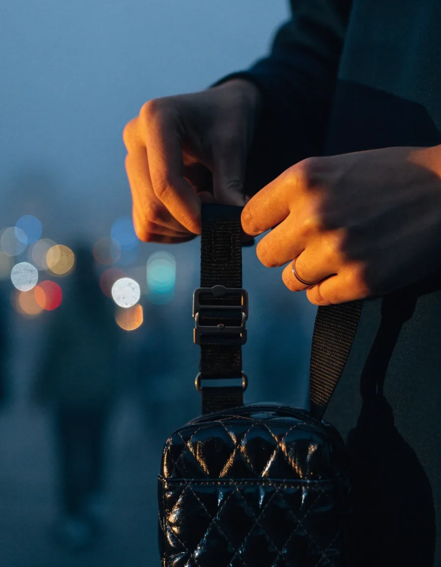 Hands adjusting the webbing strap of a small crossbody purse at hip height on a foggy evening street